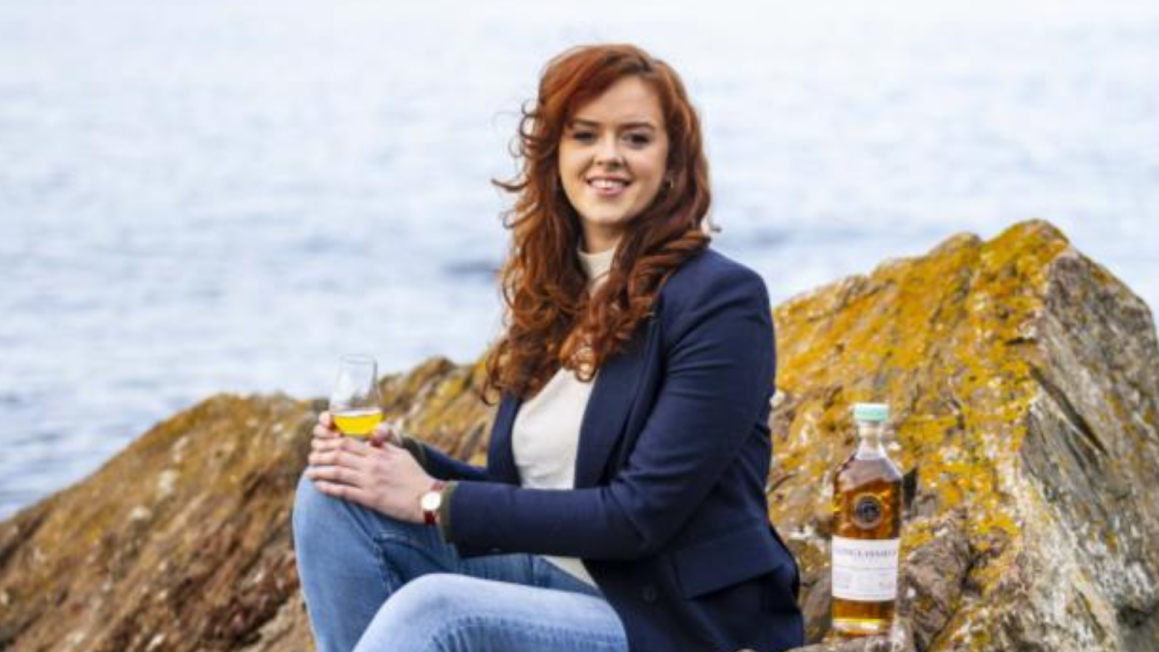 Picture of Kirsten Ainslie and a bottle of Glenglassaugh on rocks in front of water.