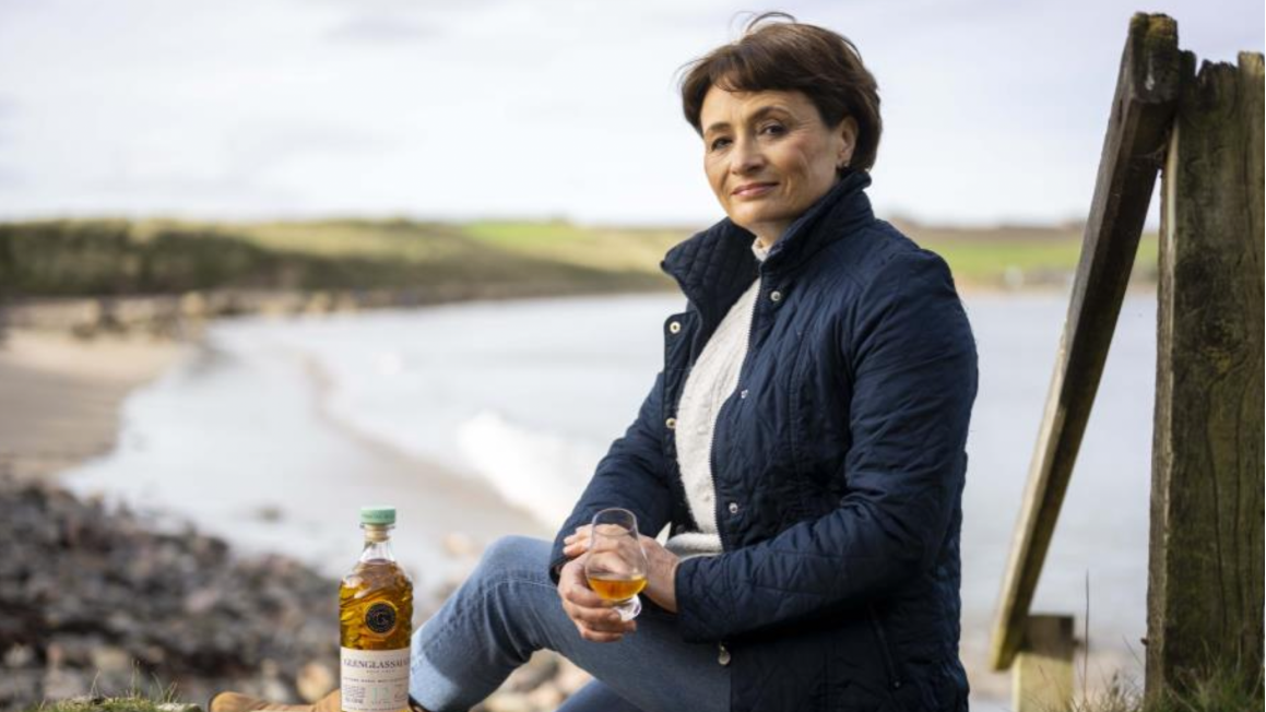 Picture of Rachel Barrie holding a glass of Glenglassaugh next to a fence with the ocean in the background.